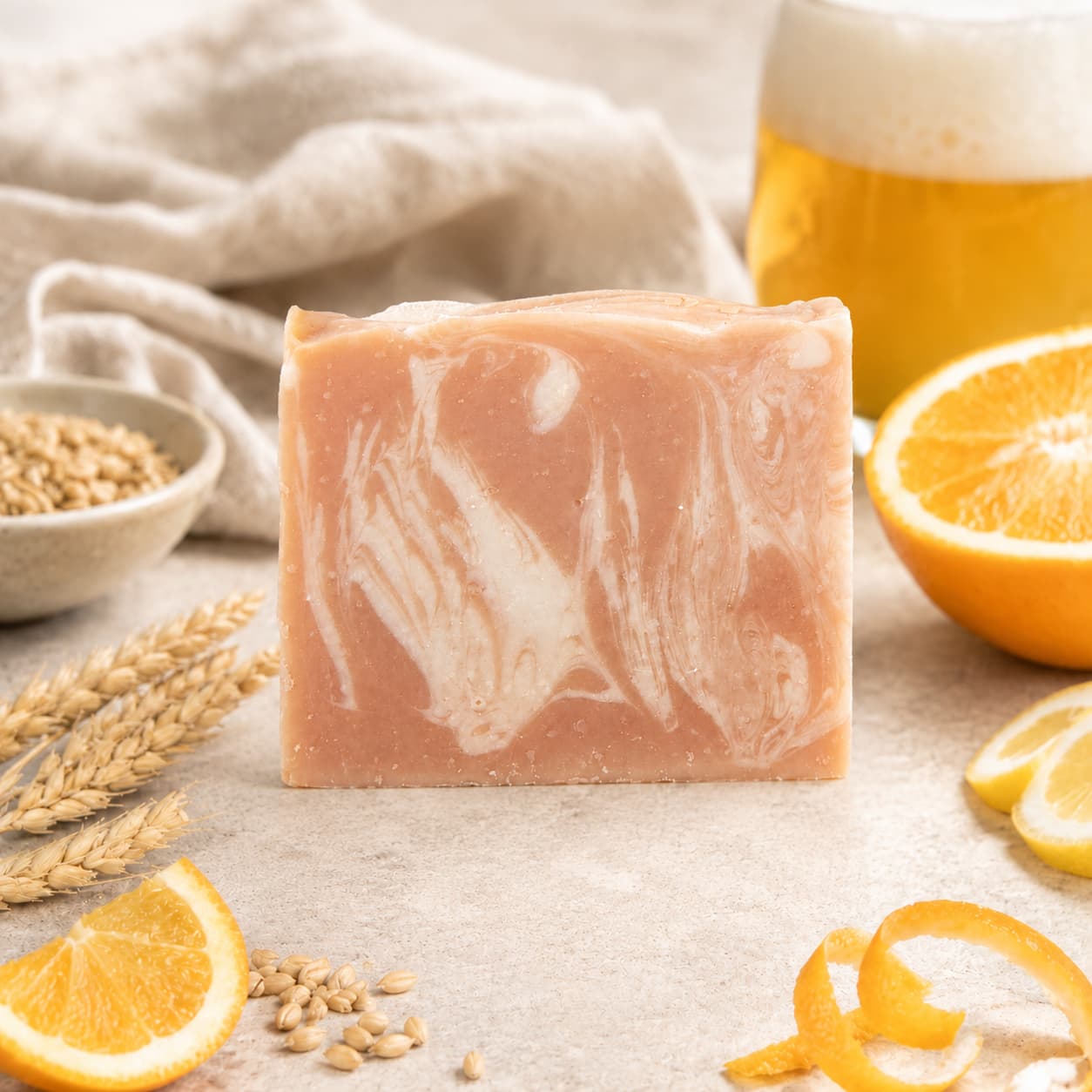 A bar of Jarhead Aromatics Belgium Wheat + Citrus Handmade Craft Beer Artisan Soap, featuring swirled white and peach colors, rests among orange slices, wheat stalks, and a frothy glass of beer on a light surface. A beige cloth is visible in the background.