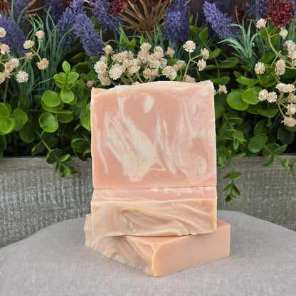 Two bars of Jarhead Aromatics Belgium Wheat + Citrus Handmade Craft Beer Artisan Soap, featuring aloe and turmeric, are stacked on a beige surface with green leaves and colorful flowers in the background.
