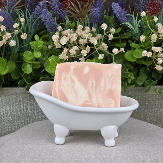 The Jarhead Aromatics Belgium Wheat + Citrus Handmade Craft Beer Soap with aloe and turmeric rests in a white clawfoot soap dish, set against green plants and purple flowers for a fresh, decorative display.