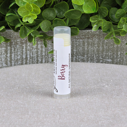 A tube of Jarhead Aromatics Tallow Lip Balm - All Natural - Assorted Flavors labeled “Berry” stands upright on a light fabric, with green plants and a gray textured planter in the background.