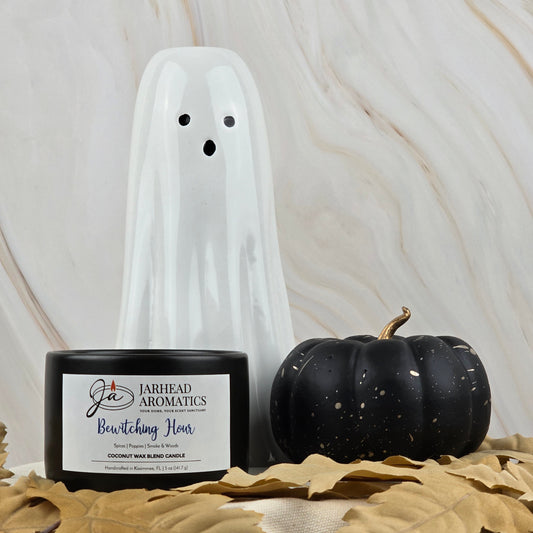 A white ceramic ghost, Jarhead Aromatics’ Bewitching Hour coconut wax blend candle with an enchanting floral scent, and a black pumpkin are arranged on beige leaves against a marble background.