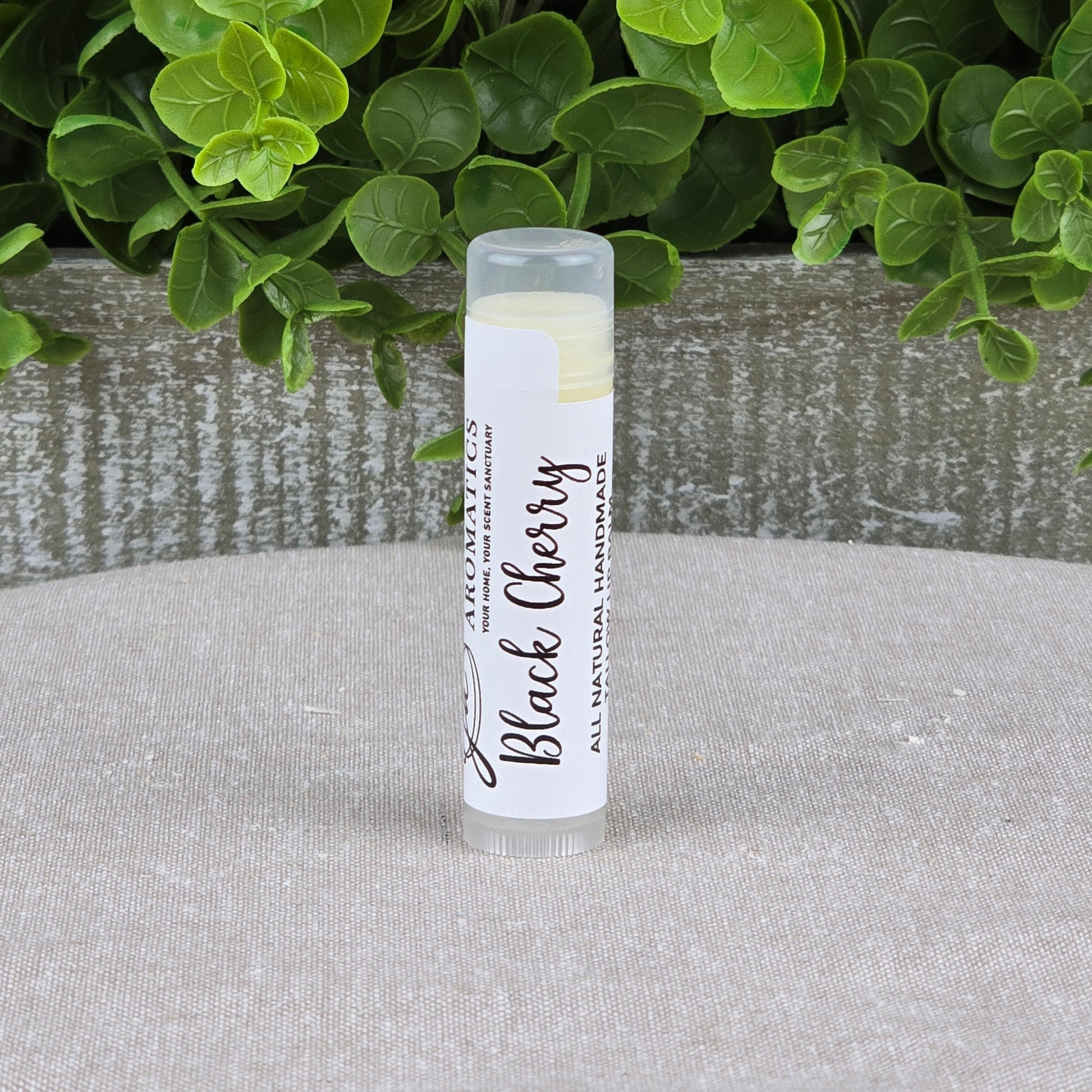 A tube of Jarhead Aromatics Tallow Lip Balm - All Natural - Assorted Flavors stands upright on a light surface, with green leafy plants and a rustic wooden container in the background, highlighting this nourishing lip balm.