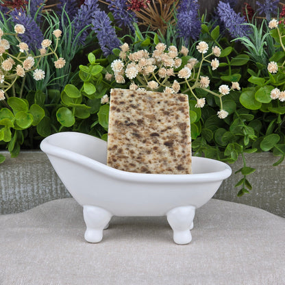 A rectangular, speckled bar of Black Soap + Lavender Handmade Artisan Soap by Jarhead Aromatics rests in a white clawfoot bathtub soap dish, with green leaves and colorful flowers in the background.