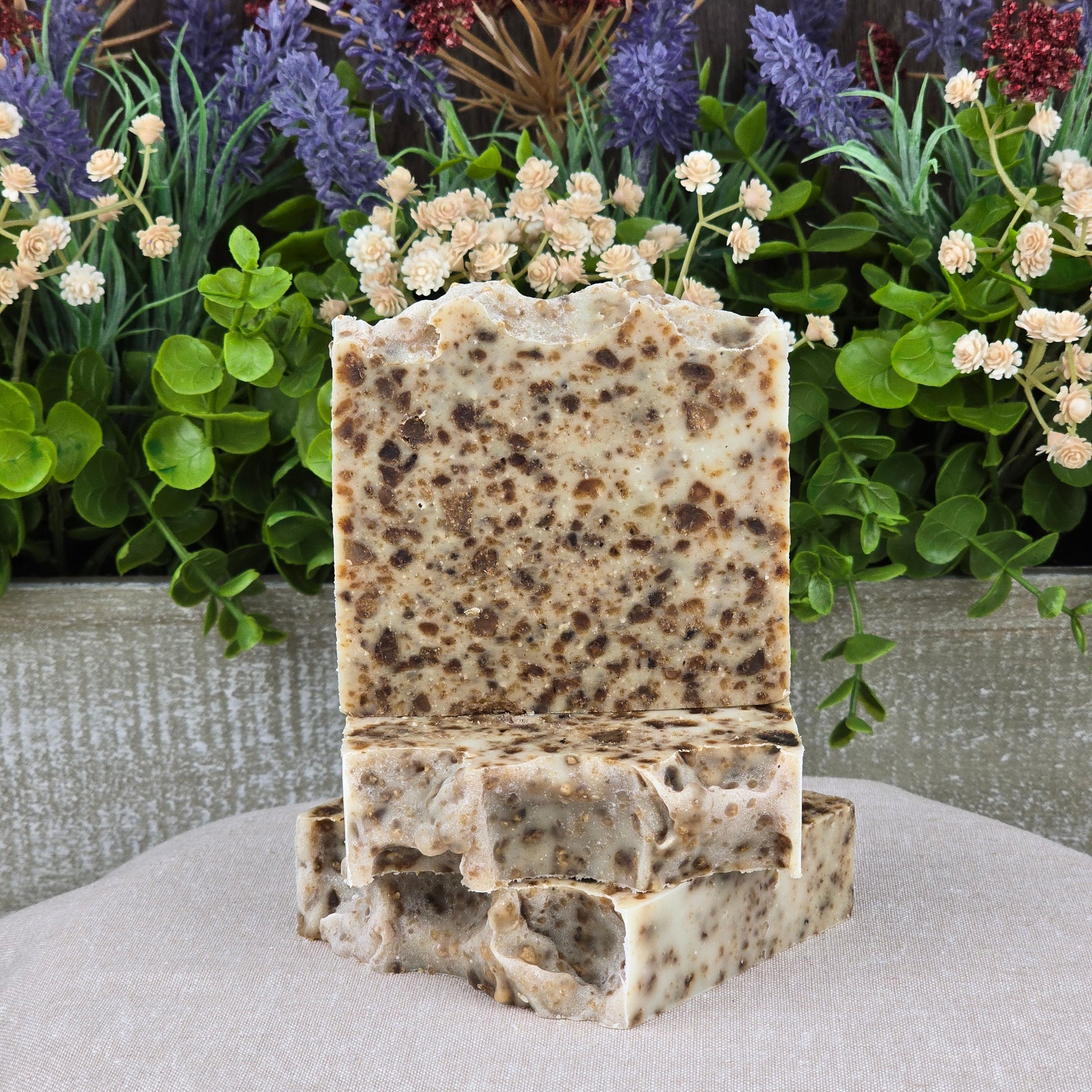Two speckled Black Soap + Lavender Handmade Natural Artisan Soap bars by Jarhead Aromatics are stacked on a beige surface, with bright green leaves and clusters of purple and cream flowers in the background.