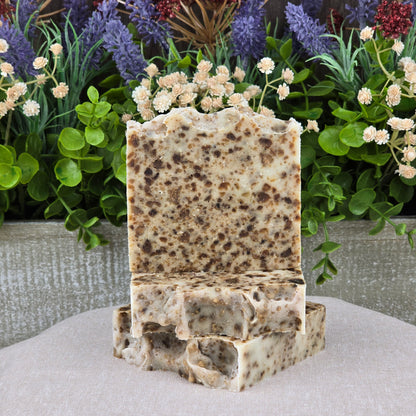 Two speckled Black Soap + Lavender Handmade Natural Artisan Soap bars by Jarhead Aromatics are stacked on a beige surface, with bright green leaves and clusters of purple and cream flowers in the background.