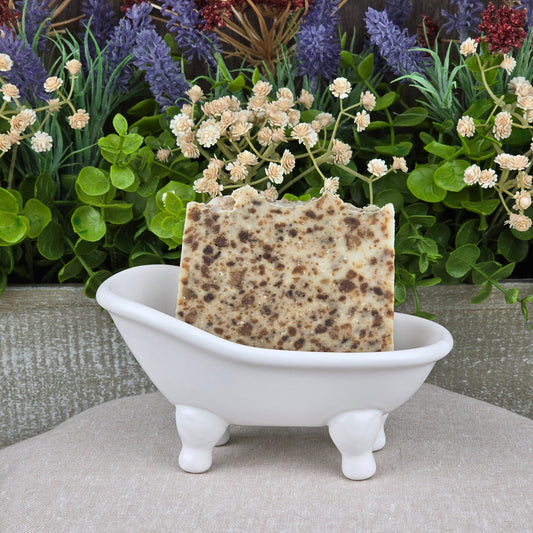 A rectangular, speckled bar of Black Soap + Lavender Handmade Natural Artisan Soap by Jarhead Aromatics sits in a small, white clawfoot bathtub soap dish, with green plants and colorful flowers in the background.