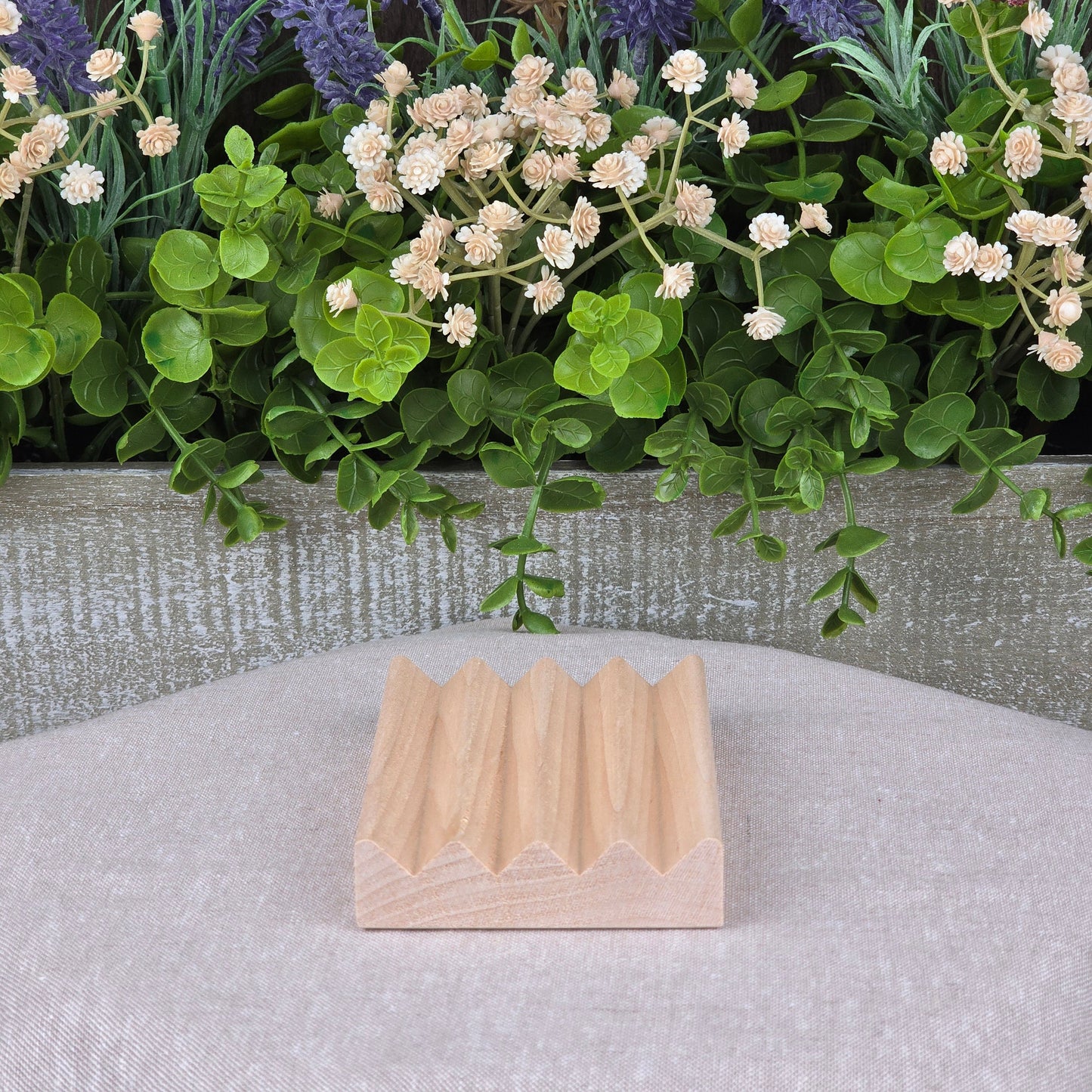 A compact, lightweight Boardwalk Style Wood Soap Dish by Jarhead Aromatics rests on beige fabric. Its zigzag top contrasts a decorative planter behind it, filled with lush green leaves and white and purple flowers.