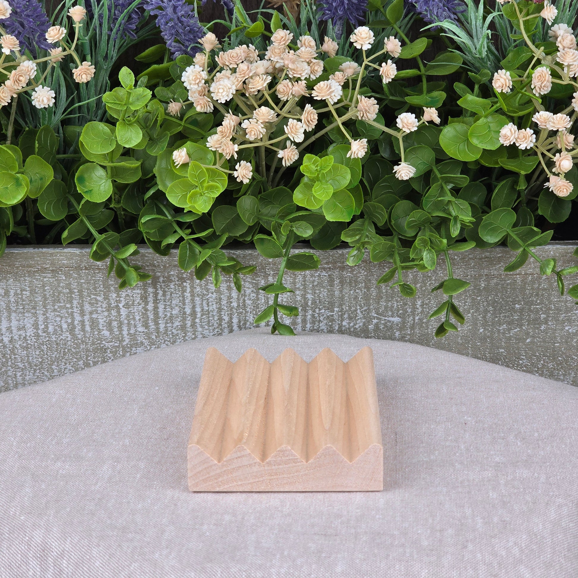A compact, lightweight Boardwalk Style Wood Soap Dish by Jarhead Aromatics rests on beige fabric. Its zigzag top contrasts a decorative planter behind it, filled with lush green leaves and white and purple flowers.