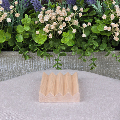 A compact, lightweight Boardwalk Style Wood Soap Dish by Jarhead Aromatics rests on beige fabric. Its zigzag top contrasts a decorative planter behind it, filled with lush green leaves and white and purple flowers.