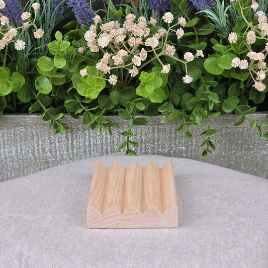 A compact, lightweight Boardwalk Style Wood Soap Dish by Jarhead Aromatics rests on beige fabric. Its zigzag top contrasts a decorative planter behind it, filled with lush green leaves and white and purple flowers.