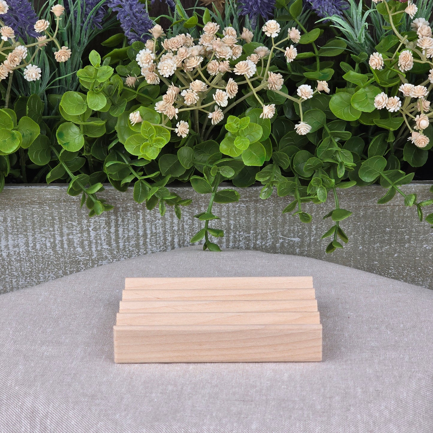 The Boardwalk Style Wood Soap Dish by Jarhead Aromatics, a handmade sustainable rectangular Red Alder soap saver, is shown on gray fabric with green leaves and white flowers in the background.
