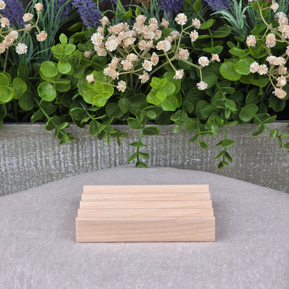 The Boardwalk Style Wood Soap Dish by Jarhead Aromatics, a handmade sustainable rectangular Red Alder soap saver, is shown on gray fabric with green leaves and white flowers in the background.