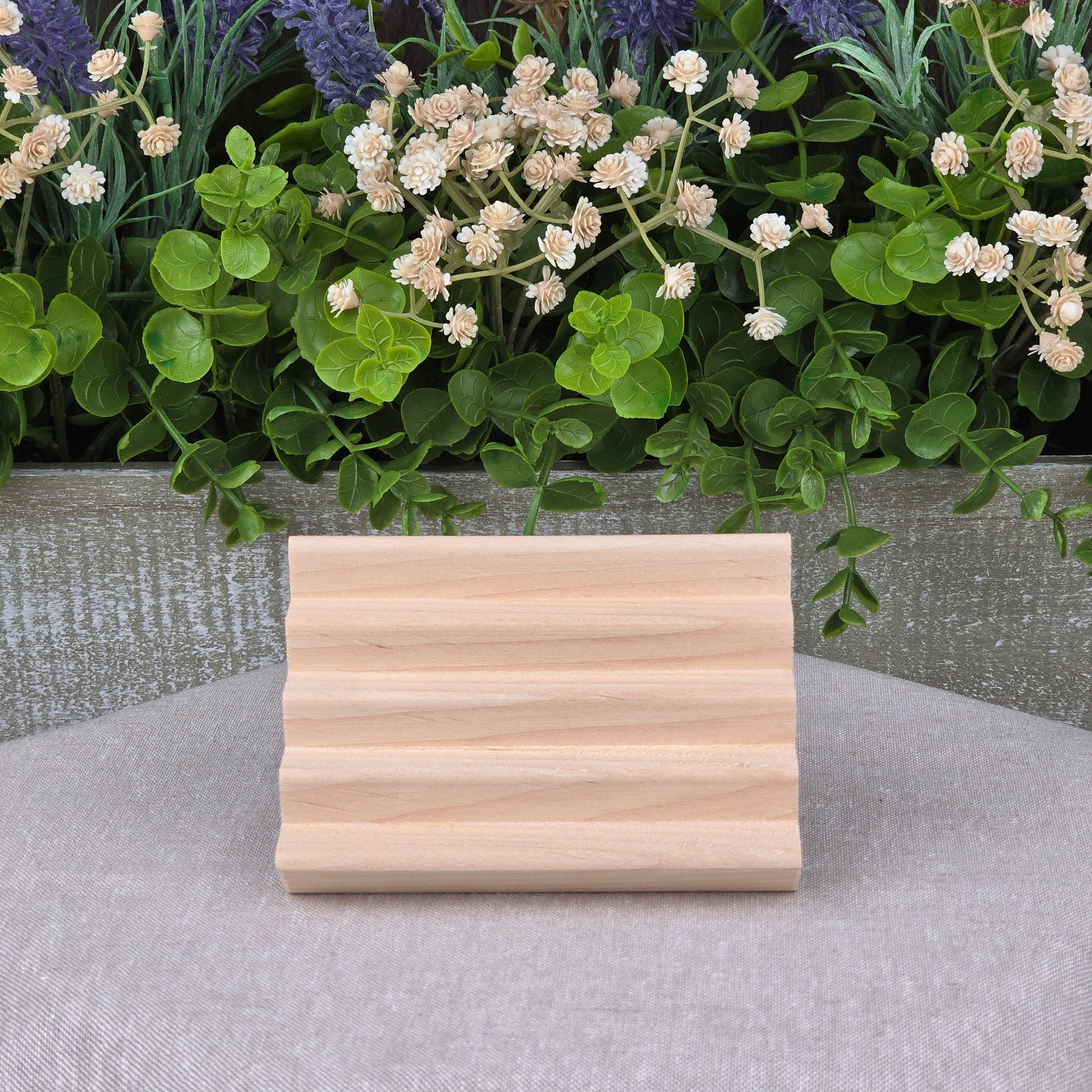 A small, lightweight Boardwalk Style Wood Soap Dish by Jarhead Aromatics sits on a gray surface, with three grooves and handmade from sustainable wood. Green and white artificial plants and purple flowers decorate the background.