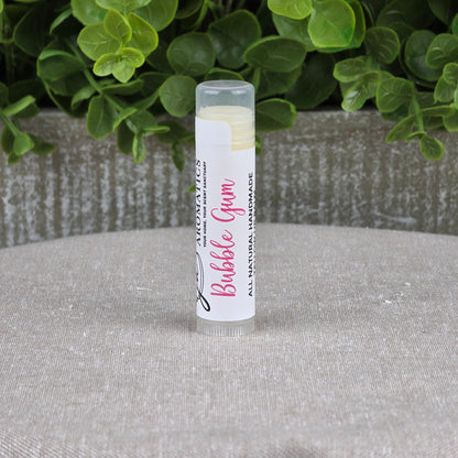 A tube of Jarhead Aromatics Tallow Lip Balm - All Natural - Assorted Flavors stands upright on a gray surface, with green leafy plants and a textured gray planter in the background.