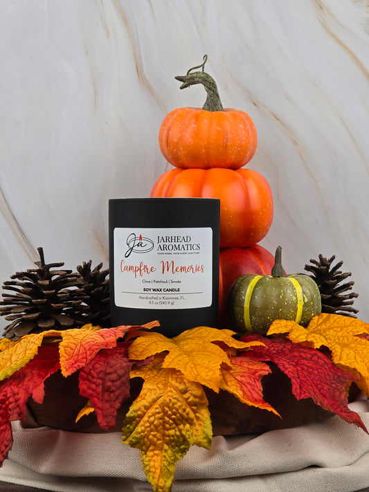 A Campfire Memories - Soy Wax Candle by Jarhead Aromatics is enveloped by pinecones, autumn leaves, and small decorative pumpkins on a marbled background. Its warm essential oil scent adds nostalgia and comfort to the cozy fall arrangement.