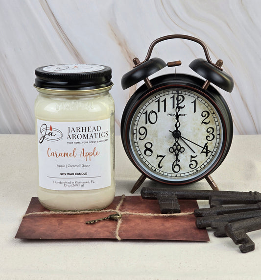 A Jarhead Aromatics Caramel Apple - Soy Wax Candle with a sweet caramel and Granny Smith apple aroma sits next to a classic black and white analog alarm clock. Below them, several rustic metal keys rest on vintage paper, all against a marble background.