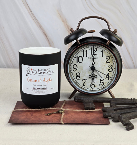 A black Caramel Apple - Soy Wax Candle by Jarhead Aromatics sits next to a vintage alarm clock. On a beige surface with antique keys and vintage paper, its aroma recalls sweet caramel drizzling over tart Granny Smith apples.