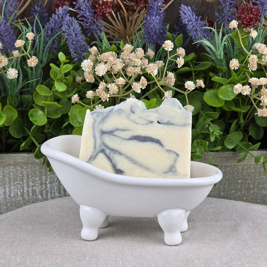 A bar of Cashmere + Santal Handmade Coconut Milk Artisan Soap by Jarhead Aromatics rests in a small, white clawfoot tub dish, with artificial green and purple flowers in the background.