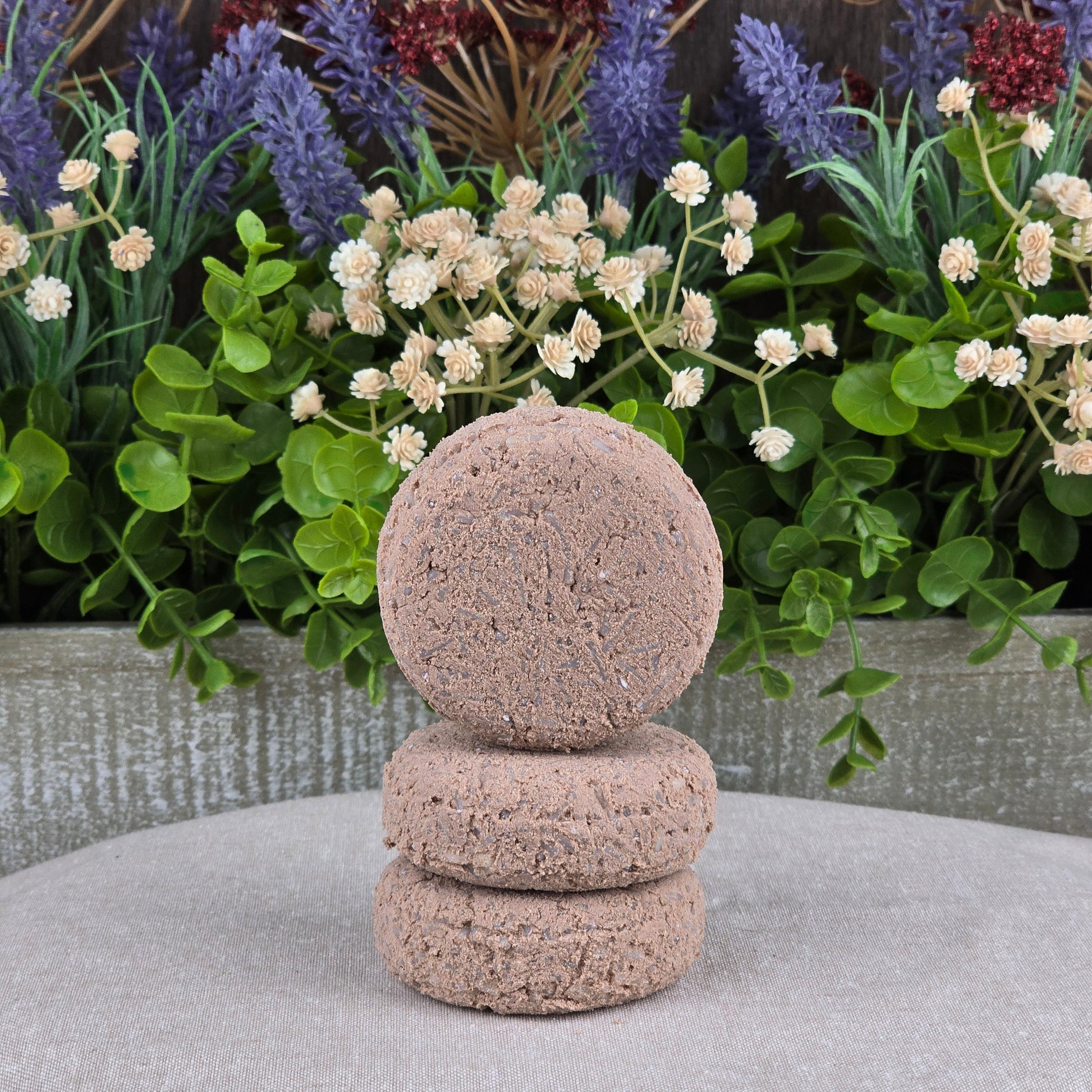 Three round Cedarwood Handmade Shampoo & Conditioner Bars by Jarhead Aromatics stacked on a gray surface, with lush greenery and purple-white artificial flowers in the background.