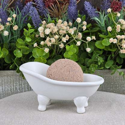 A round bath bomb rests in a white clawfoot bathtub dish on a beige surface, surrounded by greenery and flowers—perfect to pair with Jarhead Aromatics’ Cedarwood Handmade Shampoo & Conditioner Bar for a spa-like touch.