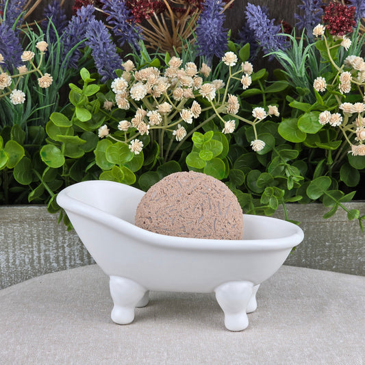 A round bath bomb rests in a white clawfoot bathtub dish on a beige surface, surrounded by greenery and flowers—perfect to pair with Jarhead Aromatics’ Cedarwood Handmade Shampoo & Conditioner Bar for a spa-like touch.
