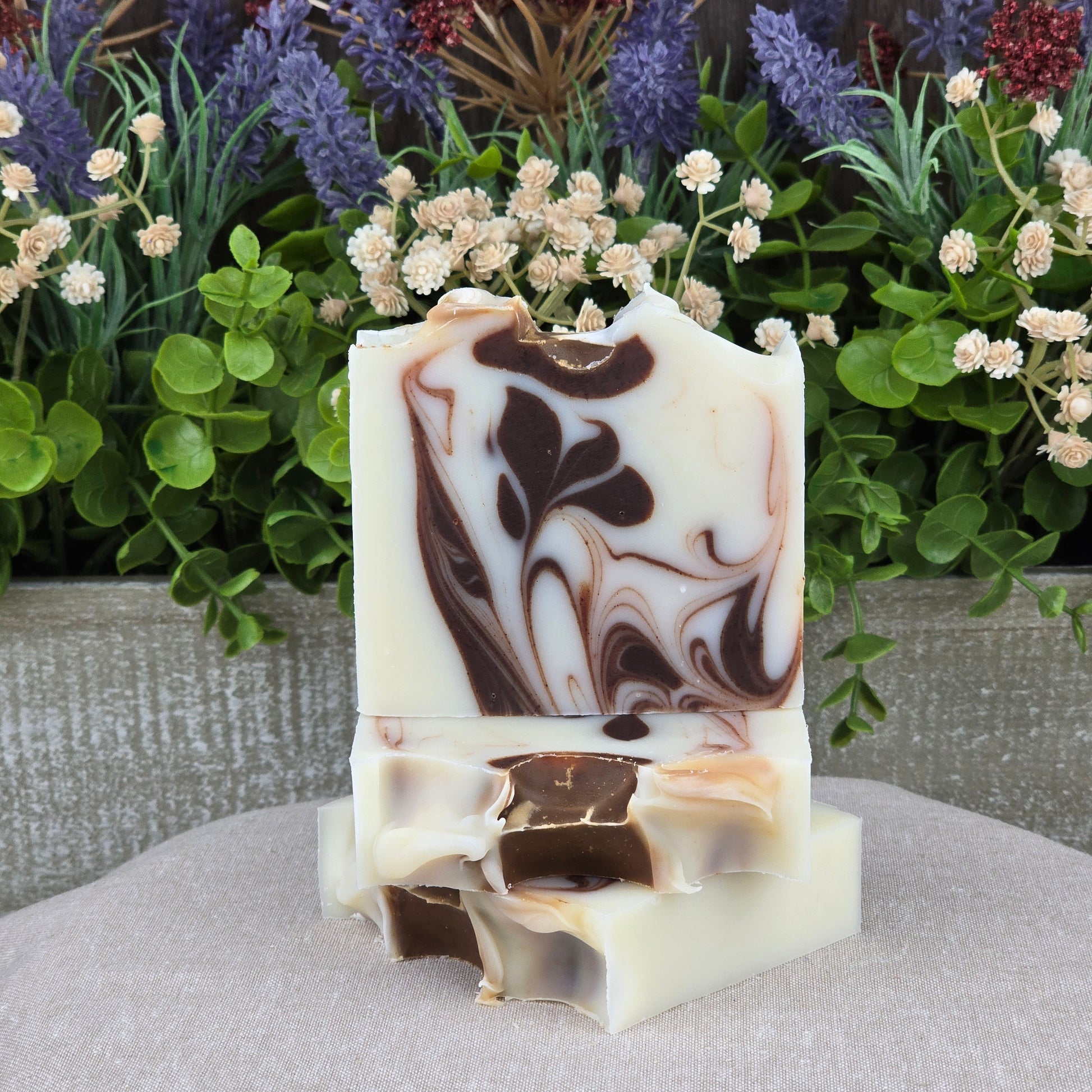 Three bars of Jarhead Aromatics Cedarwood + Cocoa Handmade Natural Tallow Artisan Soap, with brown and cream swirls, are stacked in front of lush green leaves and purple and white flowers on a neutral fabric background.