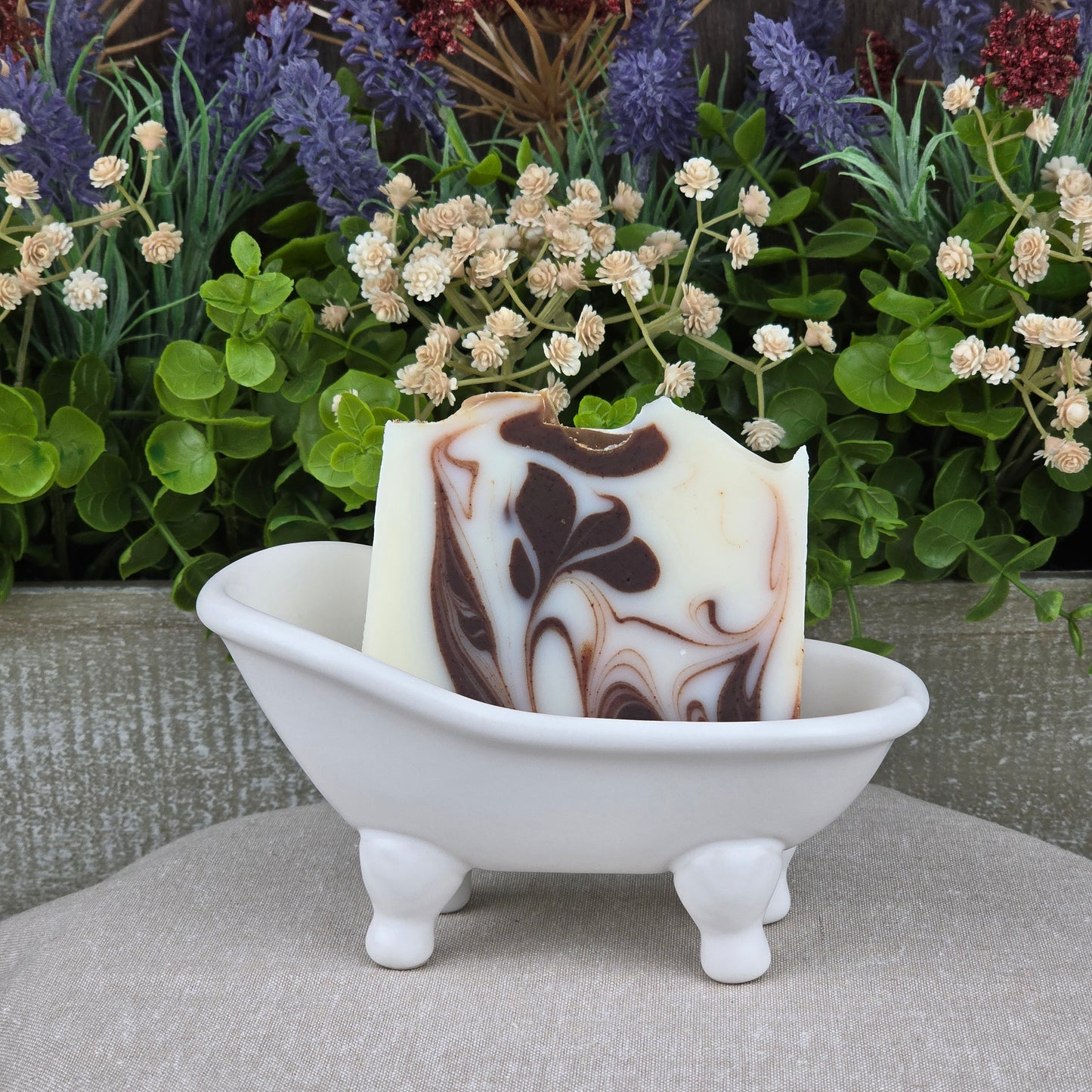 A bar of Jarhead Aromatics Cedarwood + Cocoa Handmade Natural Tallow Artisan Soap with brown and cream swirls sits in a white ceramic bathtub dish, set against a backdrop of lush green plants and purple flowers.