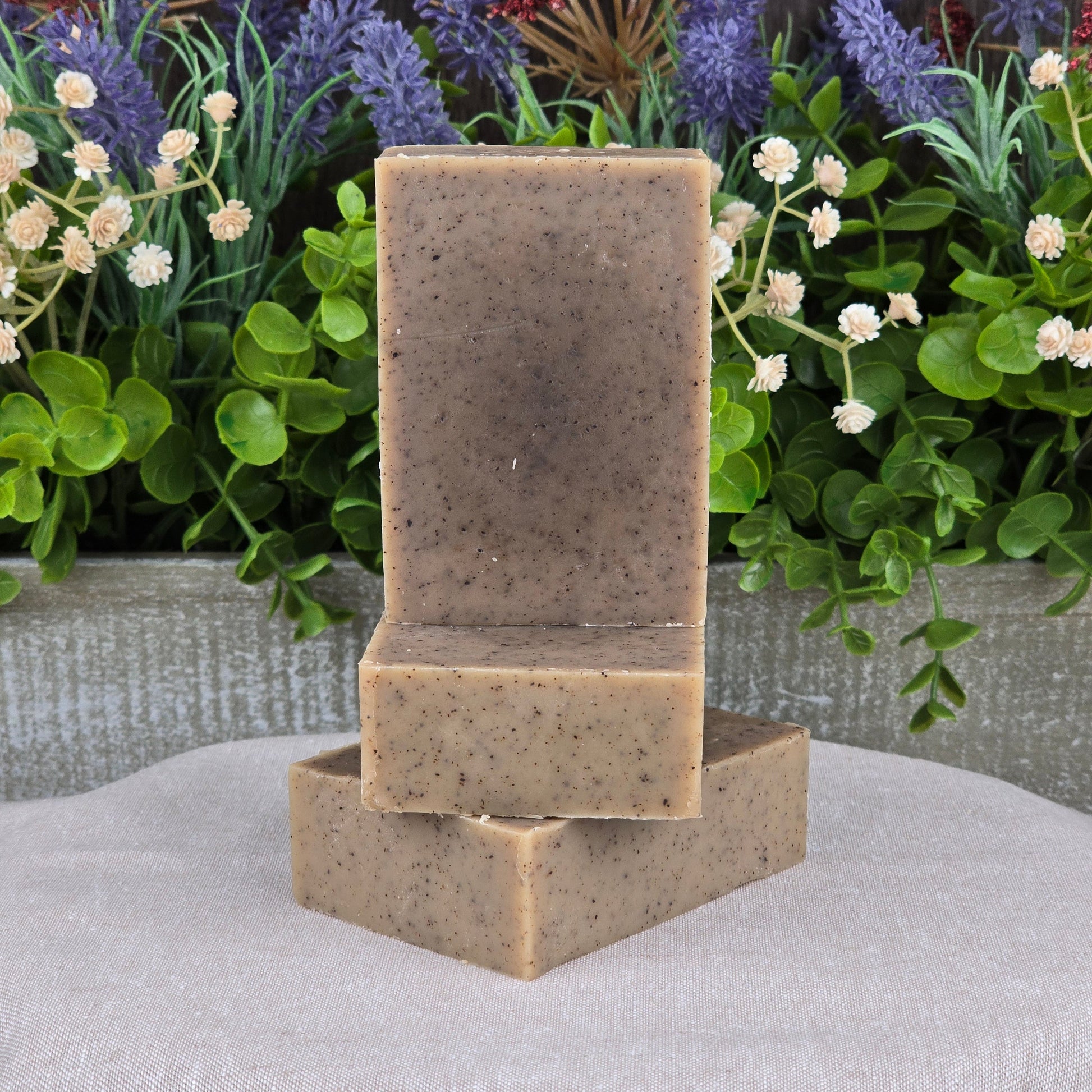 Two bars of Jarhead Aromatics Cedarwood + Coffee Handmade Soap are stacked on a neutral cloth, with green leaves and purple and white flowers in the background.