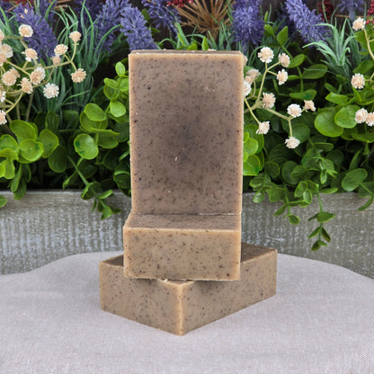 Two bars of Jarhead Aromatics Cedarwood + Coffee Handmade Soap are stacked on a neutral cloth, with green leaves and purple and white flowers in the background.