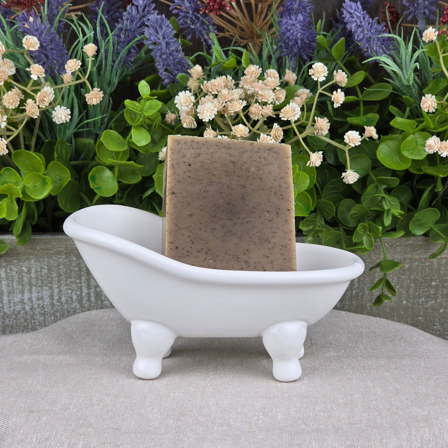 A rectangular bar of Jarhead Aromatics Cedarwood + Coffee Handmade Soap rests in a small, white clawfoot bathtub-shaped soap dish, with green plants and cream-colored flowers in the background.