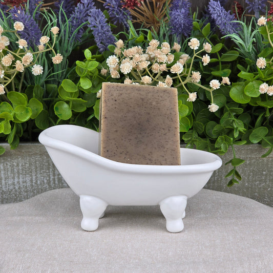 A rectangular bar of Jarhead Aromatics Cedarwood + Coffee Handmade Soap rests in a small, white clawfoot bathtub-shaped soap dish, with green plants and cream-colored flowers in the background.