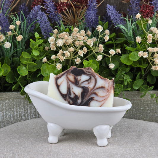 Jarhead Aromatics’ Cedarwood + Tallow Handmade Artisan Soap, featuring a brown and white marbled pattern, sits in a white clawfoot bathtub dish with green leaves and faux white and purple flowers as a charming backdrop.