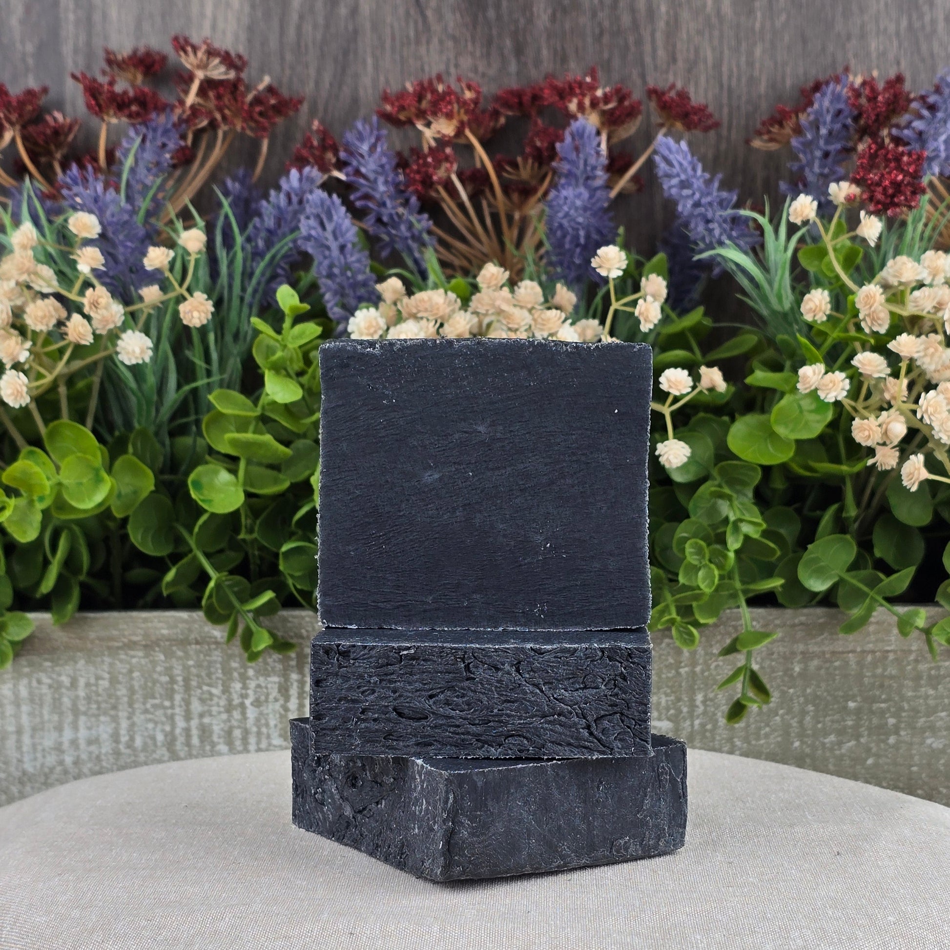 Jarhead Aromatics Charcoal Handmade Soap—an all-natural black rectangular bar—is displayed on two rough stones atop beige cloth, with green leaves and colorful artificial flowers in the background; ideal for natural skincare.