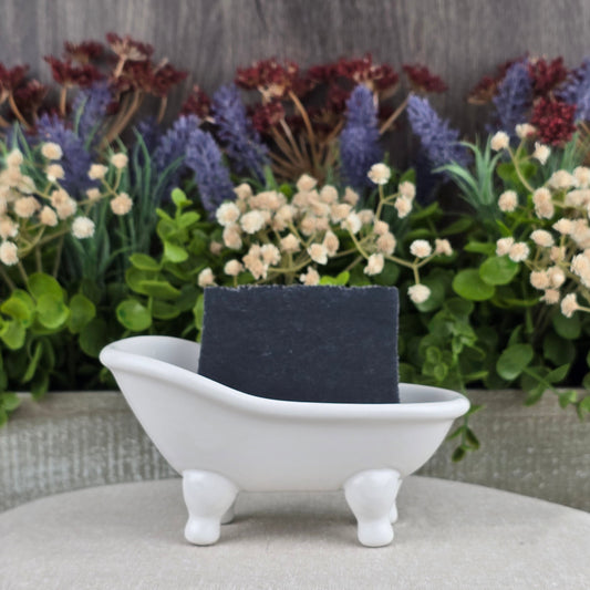 A bar of Charcoal Handmade Soap - All Natural by Jarhead Aromatics sits in a white clawfoot bathtub soap dish, with artificial flowers and greenery arranged decoratively in the background.