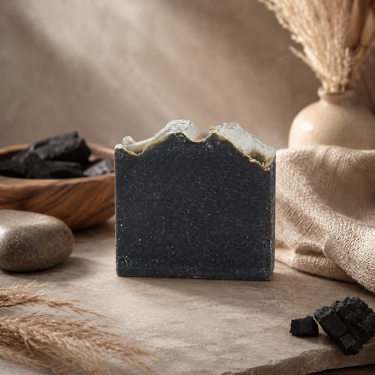 A bar of Jarhead Aromatics’ Charcoal + Tonka Handmade Artisan Soap, featuring a textured top, rests on a stone surface amidst dried grasses, stones, pieces of activated charcoal, and a beige vase in a serene neutral-toned setting.
