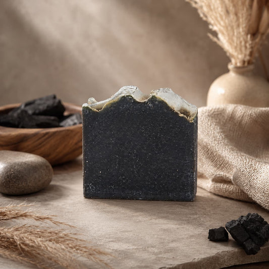 A bar of Jarhead Aromatics’ Charcoal + Tonka Handmade Artisan Soap, featuring a textured top, rests on a stone surface amidst dried grasses, stones, pieces of activated charcoal, and a beige vase in a serene neutral-toned setting.