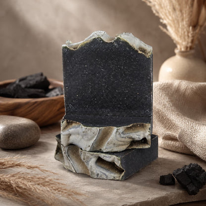 Two Charcoal + Tonka Handmade Artisan Soaps from Jarhead Aromatics, featuring uneven, wavy tops and striking marbled patterns in dark gray, are stacked atop a stone surface. In the background, dried botanicals, pieces of activated charcoal, and a wooden bowl enhance the scene’s rustic, natural appeal.