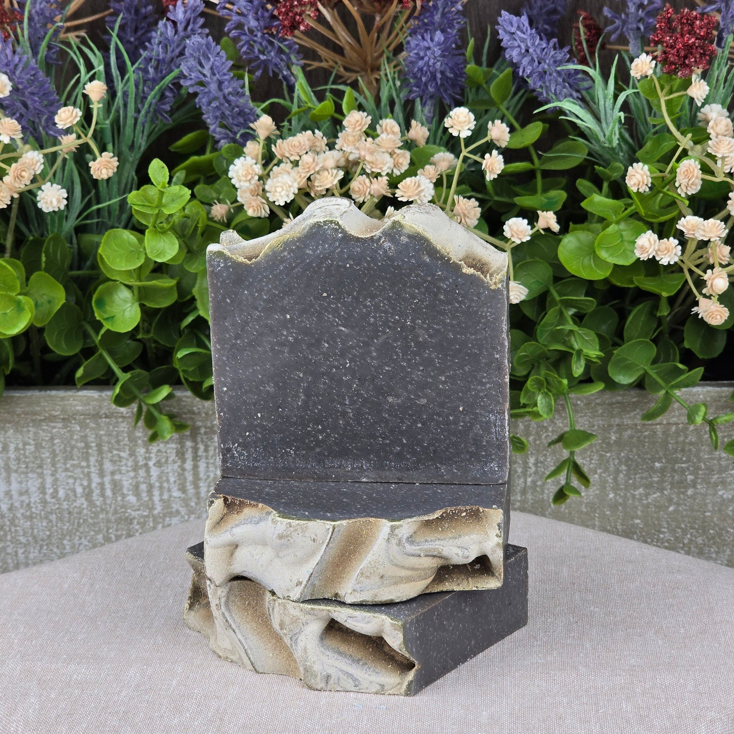Two Charcoal + Tonka Handmade Artisan Soap bars from Jarhead Aromatics, featuring marbled beige and dark gray with activated charcoal and a wavy textured top, are stacked before assorted artificial flowers and greenery.