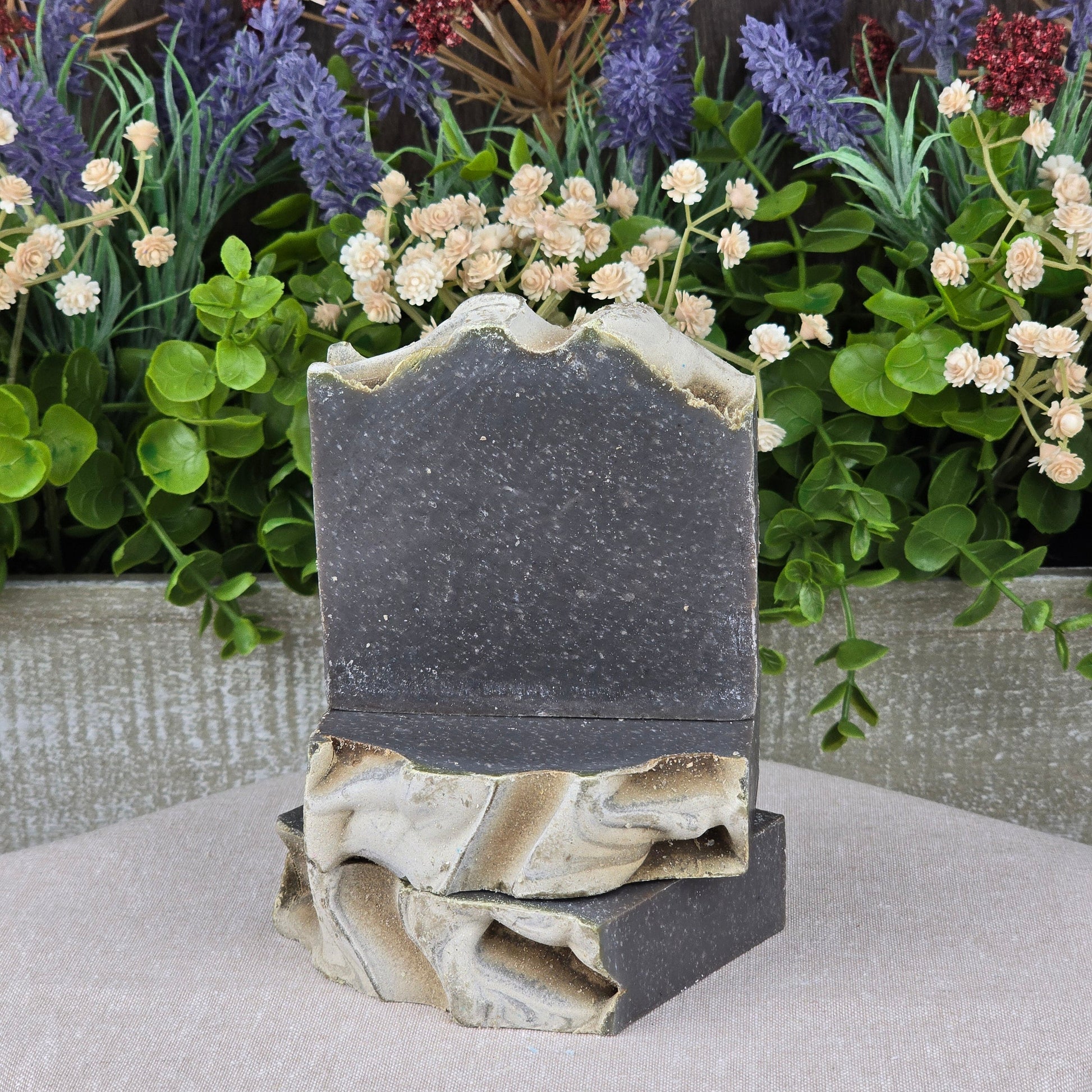 Two Charcoal + Tonka Handmade Artisan Soap bars from Jarhead Aromatics, featuring marbled beige and dark gray with activated charcoal and a wavy textured top, are stacked before assorted artificial flowers and greenery.