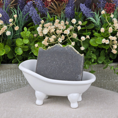 A bar of Jarhead Aromatics Charcoal + Tonka Handmade Artisan Soap sits in a small white ceramic bathtub-shaped dish, with faux green leaves and purple and cream flowers in the background.