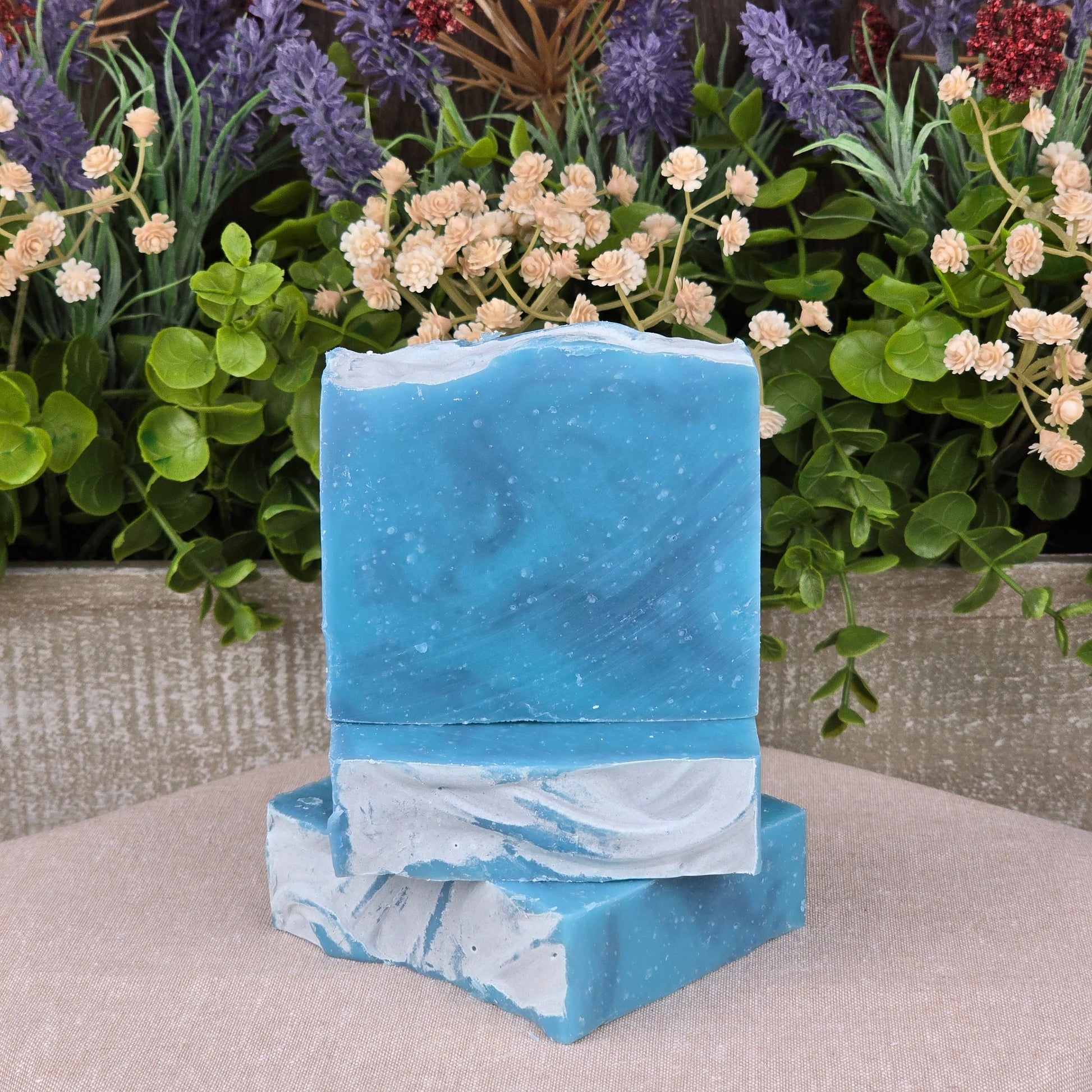 Two Citron + Cedarwood Handmade Coconut Milk Artisan Soaps from Jarhead Aromatics, blue and white marbled, are stacked in front of artificial green foliage and vibrant purple, red, and white flowers on a wooden background.
