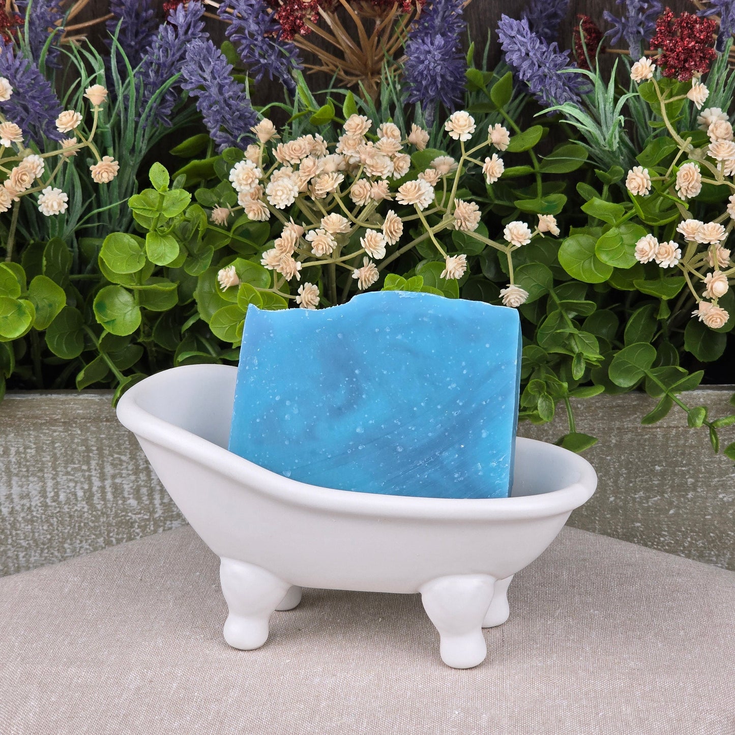 A bar of Citron + Cedarwood Handmade Coconut Milk Artisan Soap by Jarhead Aromatics sits in a small white clawfoot bathtub soap dish, with green and purple artificial flowers and plants in the background.