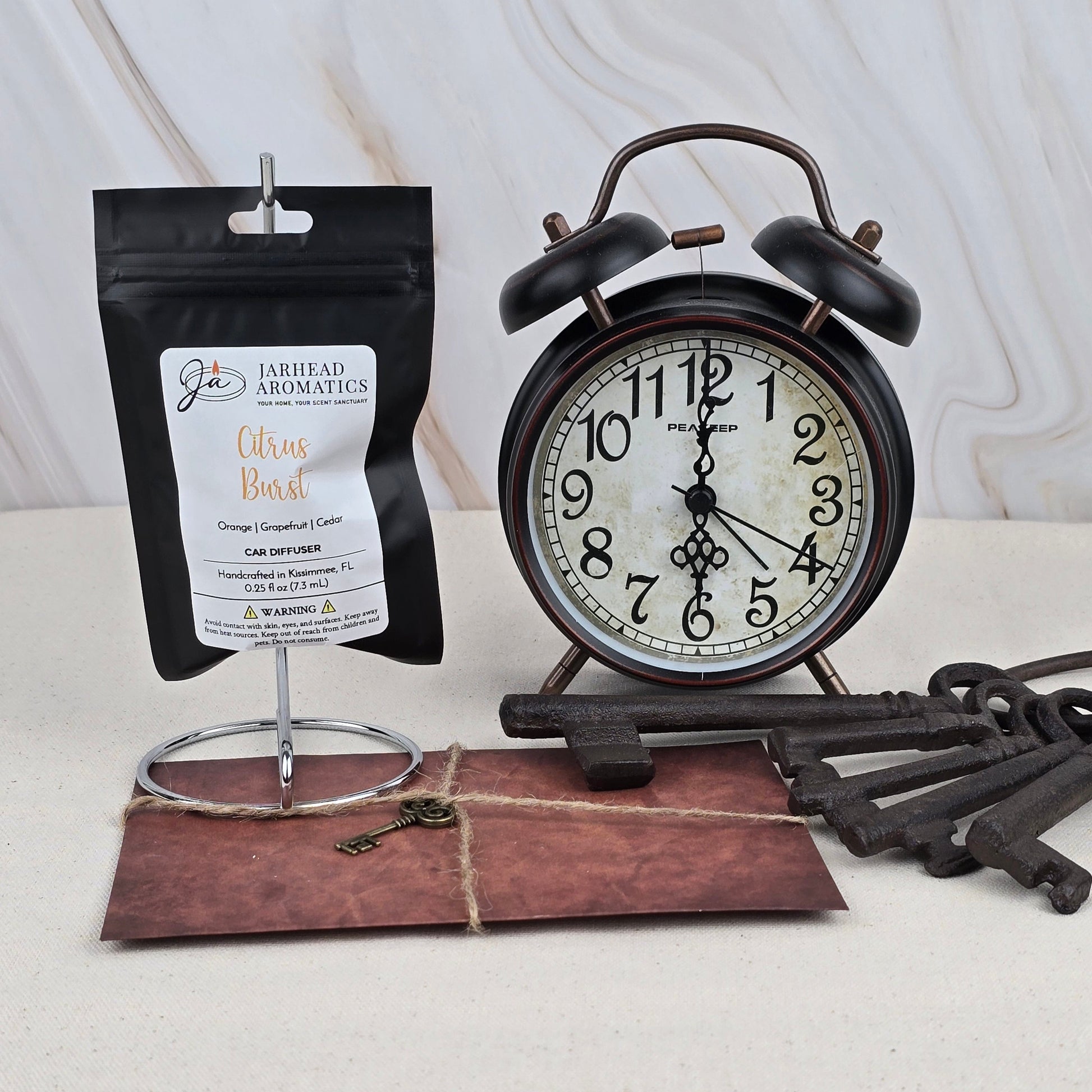 A black vintage alarm clock, Jarhead Aromatics’ Citrus Burst - Car Diffuser, an old-fashioned key on a brown envelope, and several large iron keys are arranged on a cream surface with a light marble background.