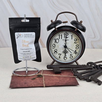 A black vintage alarm clock, Jarhead Aromatics’ Citrus Burst - Car Diffuser, an old-fashioned key on a brown envelope, and several large iron keys are arranged on a cream surface with a light marble background.
