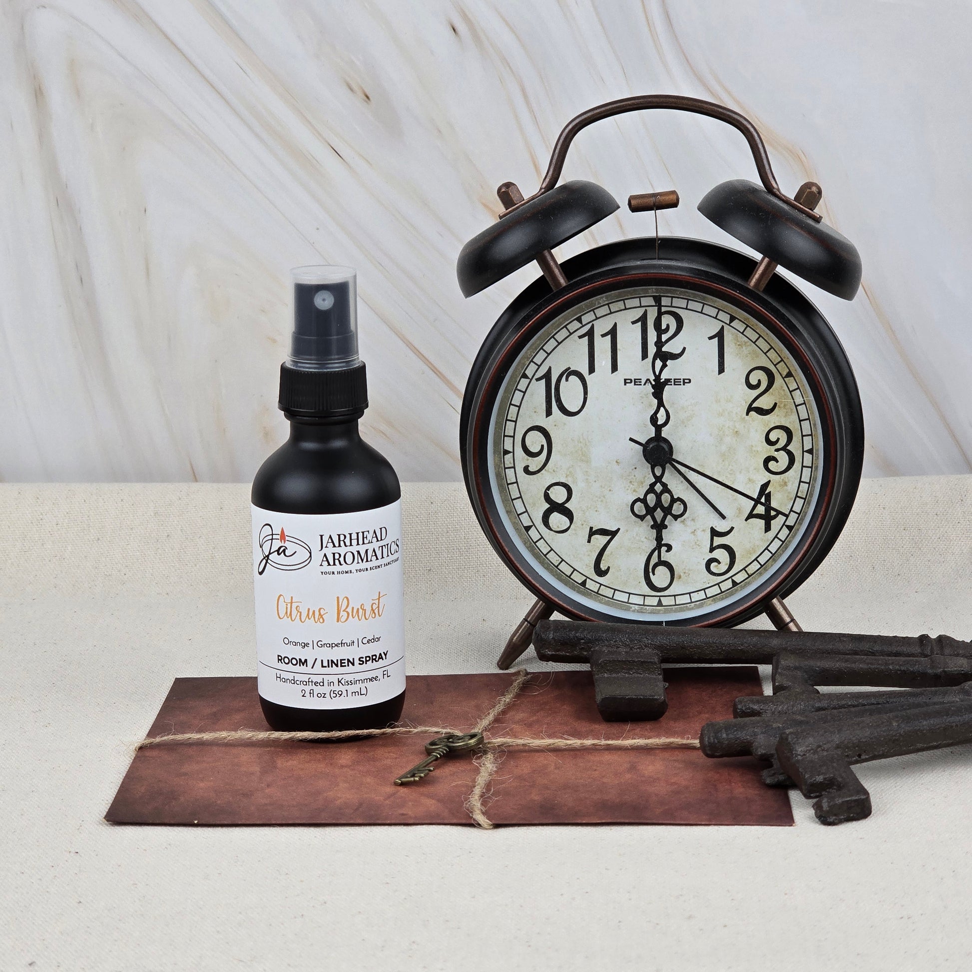 A bottle of Jarhead Aromatics Citrus Burst Room & Linen Spray | Orange, Mango & Peach Home Fragrance Spray sits beside a vintage black alarm clock, three large metal keys, a small key, and a brown envelope on a light surface.