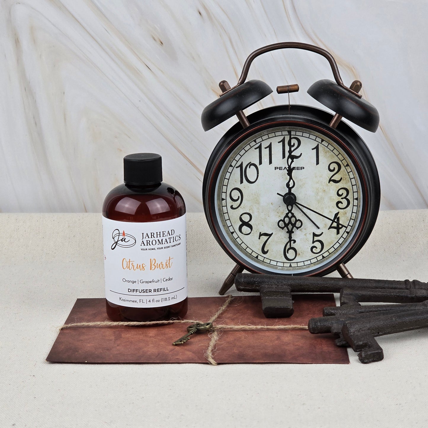 A vintage alarm clock, Jarhead Aromatics Citrus Burst Diffuser Refill | Orange, Mango & Grapefruit Home Fragrance Oil Refill, large decorative metal keys, and a small key sit on a brown leather mat against a light marble background.
