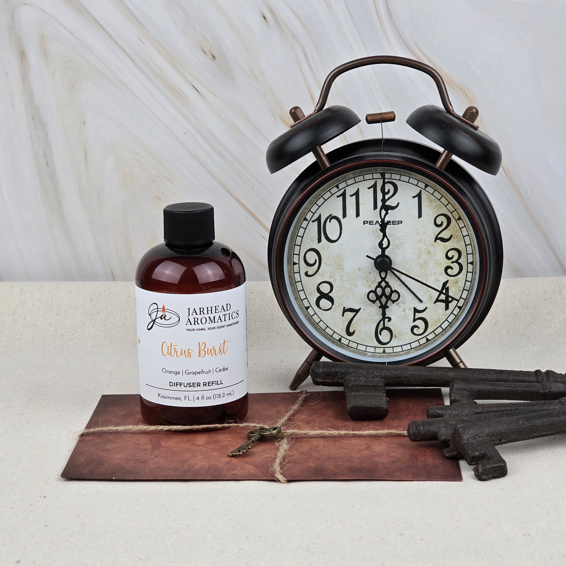 A vintage alarm clock, Jarhead Aromatics Citrus Burst Diffuser Refill | Orange, Mango & Grapefruit Home Fragrance Oil Refill, large decorative metal keys, and a small key sit on a brown leather mat against a light marble background.