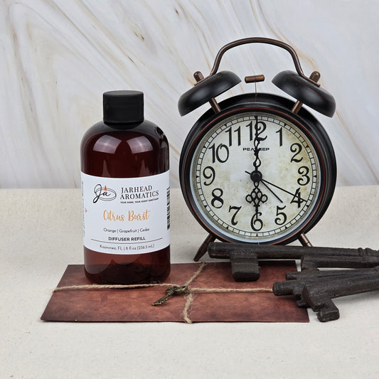 A Jarhead Aromatics Citrus Burst Diffuser Refill | Orange, Mango & Grapefruit sits by a black vintage alarm clock and three antique keys on a brown mat, its fruity aroma adding vitality to the light marble backdrop.