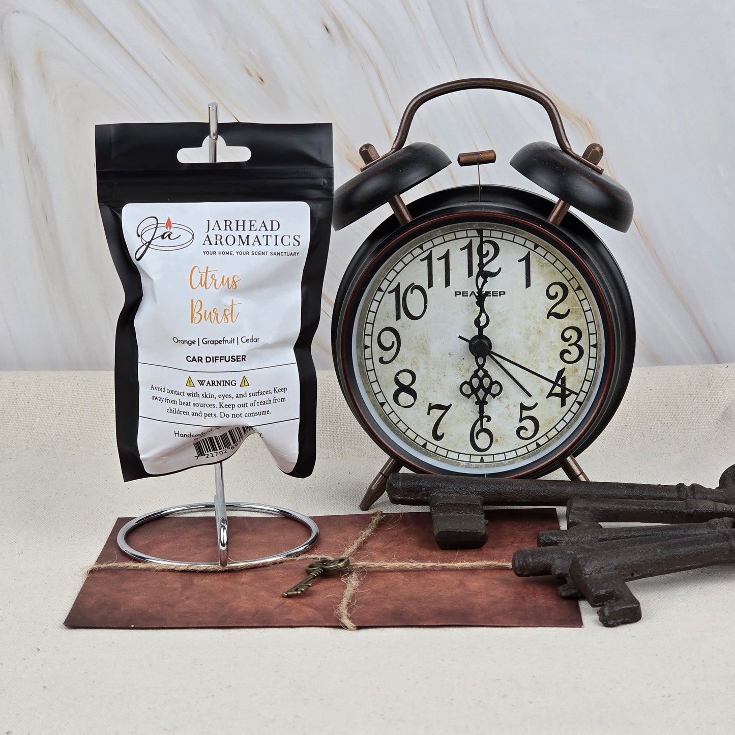 A black alarm clock showing 10:12 sits by a Jarhead Aromatics Citrus Burst Car Diffuser | Bright Citrus & Peach Car Air Freshener on a metal stand. Large vintage keys lie on brown leather below, with a marble background.