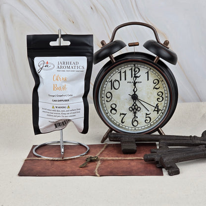 A black alarm clock showing 10:12 sits by a Jarhead Aromatics Citrus Burst Car Diffuser | Bright Citrus & Peach Car Air Freshener on a metal stand. Large vintage keys lie on brown leather below, with a marble background.